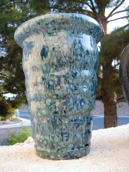 A tall vase with tapers that looks wavy for the length of the vase. The glaze is green with blue speckles.
