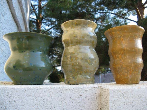 Three ceramic vases on a stone pedestal. The first vase on the left is green and has a tapered middle, the second vase is yellowish and also has a taper in the middle and is taller than the green vase, the third vase is orange and has multiple tapers and is about the same size as the green vase. All vases have a speckled look.